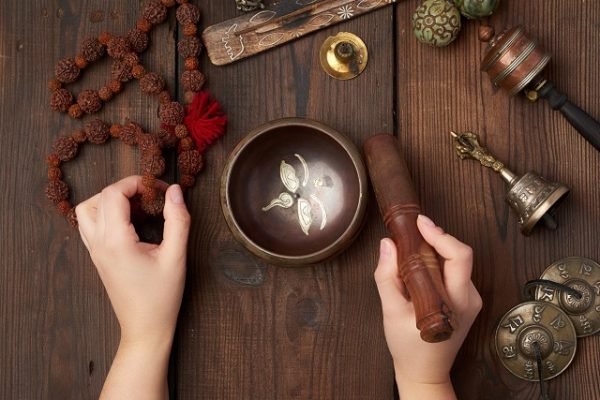 female hands hold a copper Tibetan singing bowl and a wooden stick, on the wooden table are items for alternative medicine, sound massage and meditation