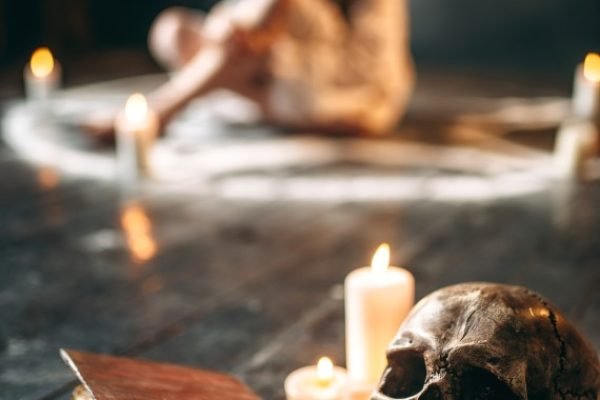 Human skull and old spellbook on black wooden floor, female person in white shirt sitting in pentagram circle with candles on background. Dark magic, occultism and exorcism